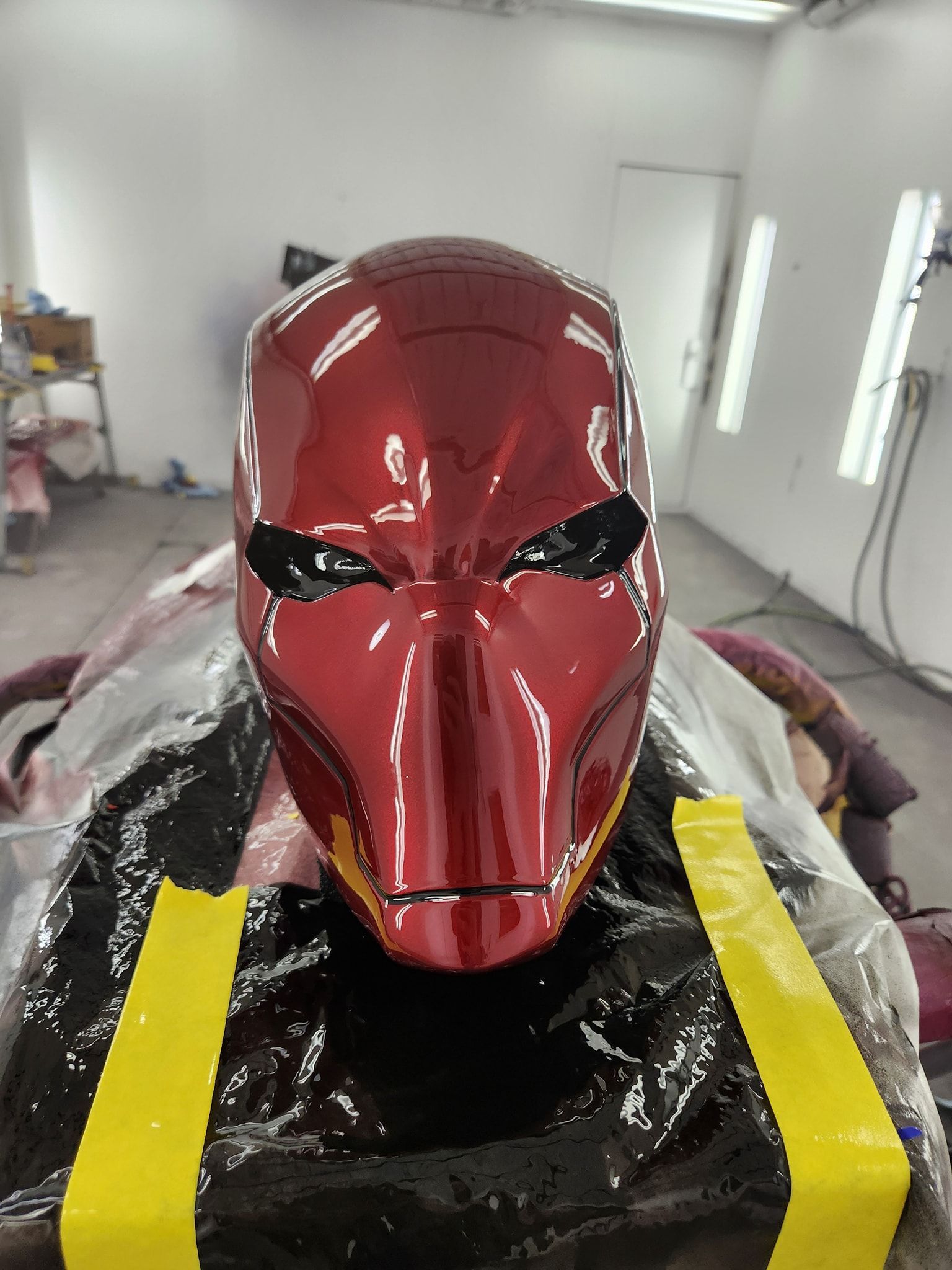 Glossy red masked sculpture on a workbench in a white paint booth, wrapped in plastic and tape.