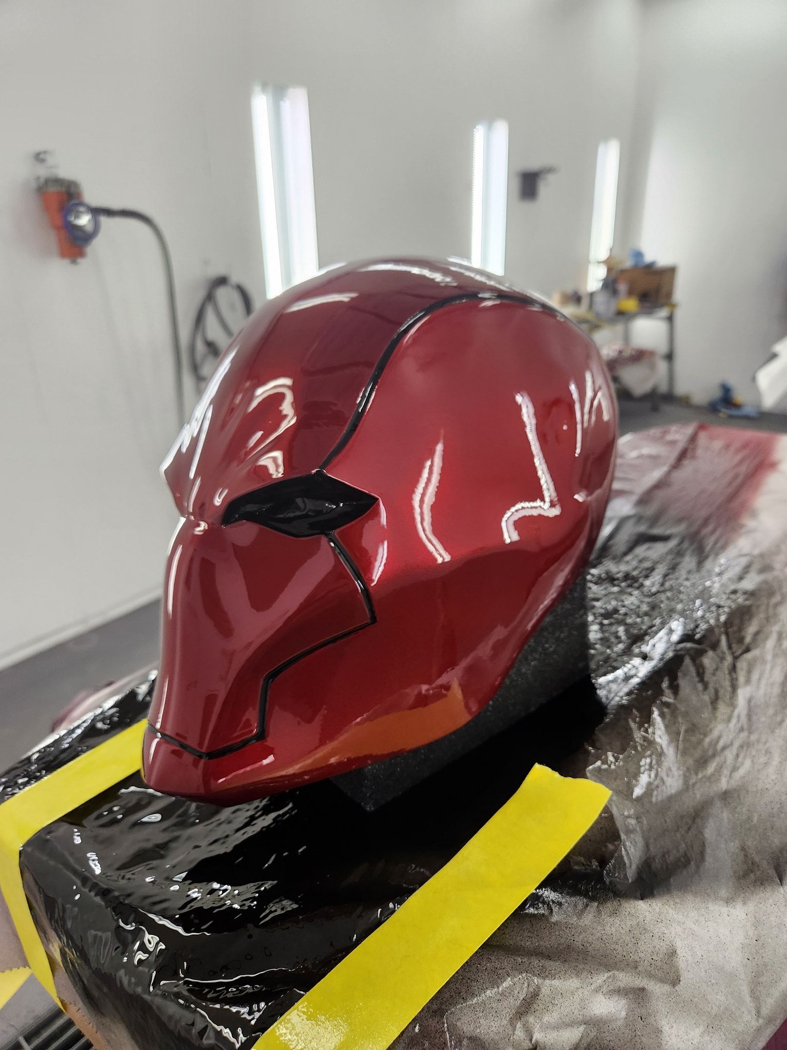 Glossy red helmet-like mask on a workbench in a workshop, with black details and yellow tape