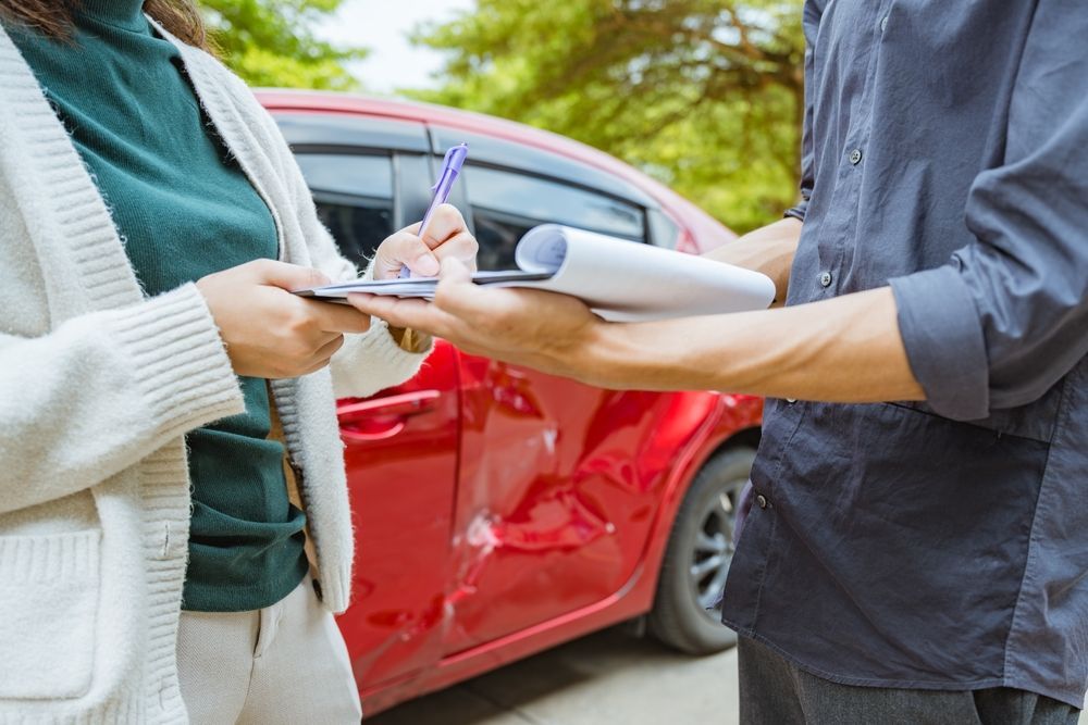 Two people exchanging insurance documents beside a red car with visible side damage.