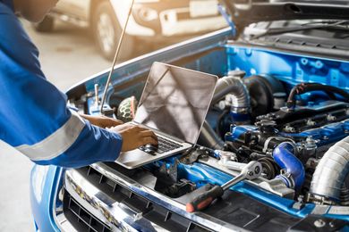 Mechanic using a laptop to diagnose a blue car’s open engine bay in a garage Mechanic using a laptop to diagnose a blue car’s open engine bay in a garage