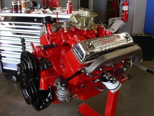 Red classic car engine on a stand in a garage, with chrome valve covers and black pulleys.