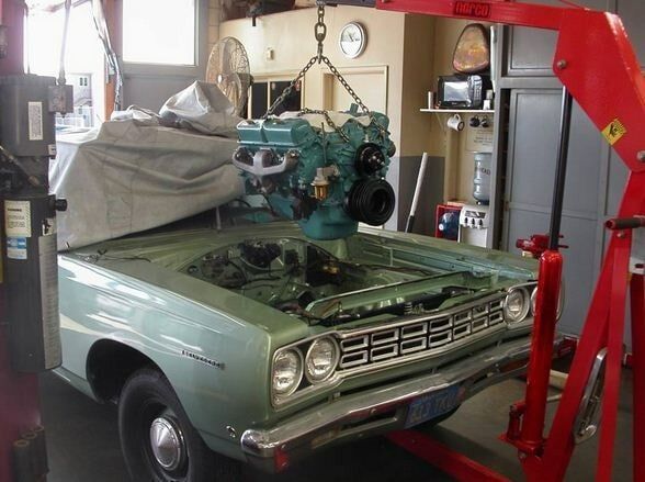 Green vintage car being lifted in a garage by a red engine hoist, with a detached engine suspended above it