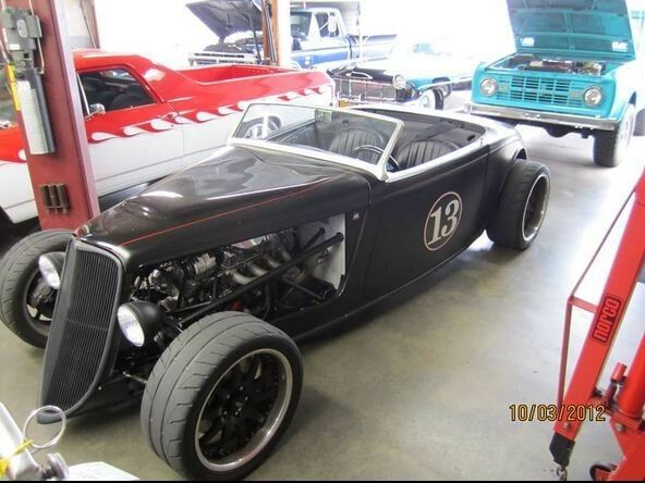 Black vintage hot rod with number 13 in a garage, with a red car and blue truck in the background