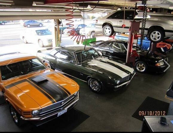 Classic cars parked in a garage, including an orange muscle car and a black convertible with white stripes.