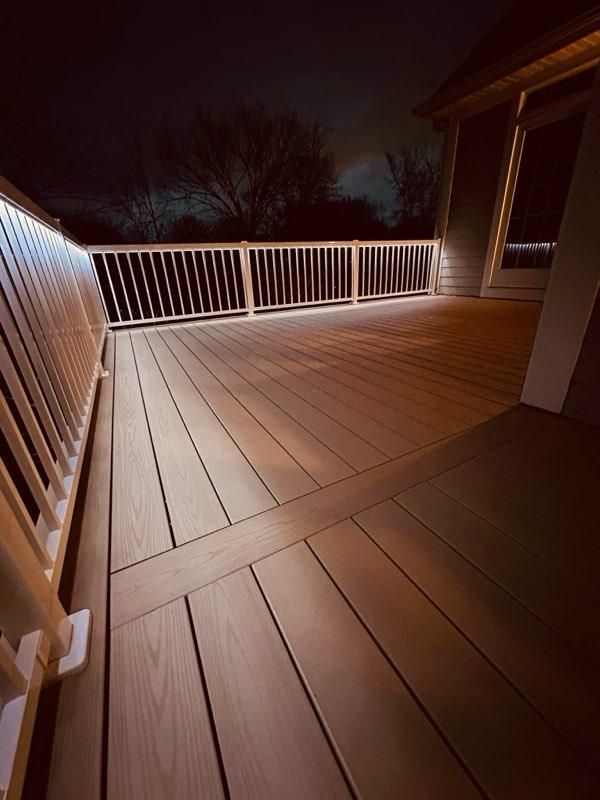 Lit wooden deck at night with white railing, glowing lights, and a glimpse of a building.