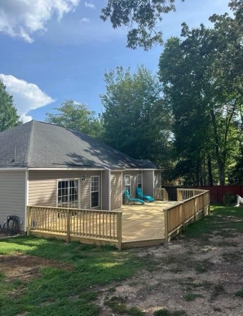 Backyard view of a home with a wooden deck. Two blue lounge chairs are on the deck. Lush green trees surround the house.