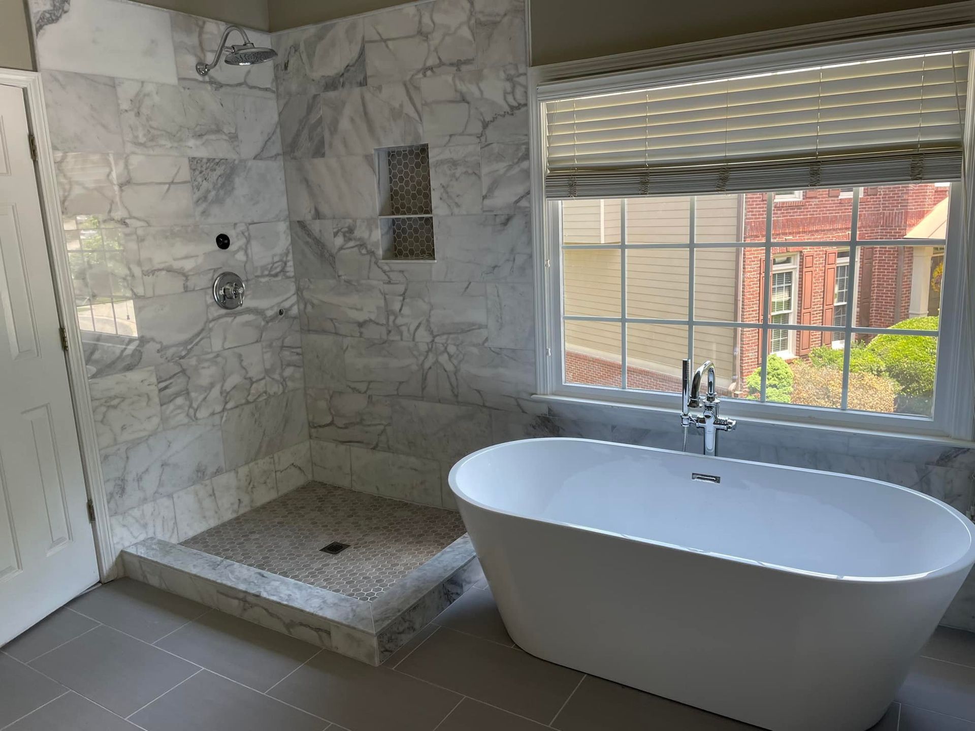 A modern bathroom with a white tub, marble shower, and a window with blinds.