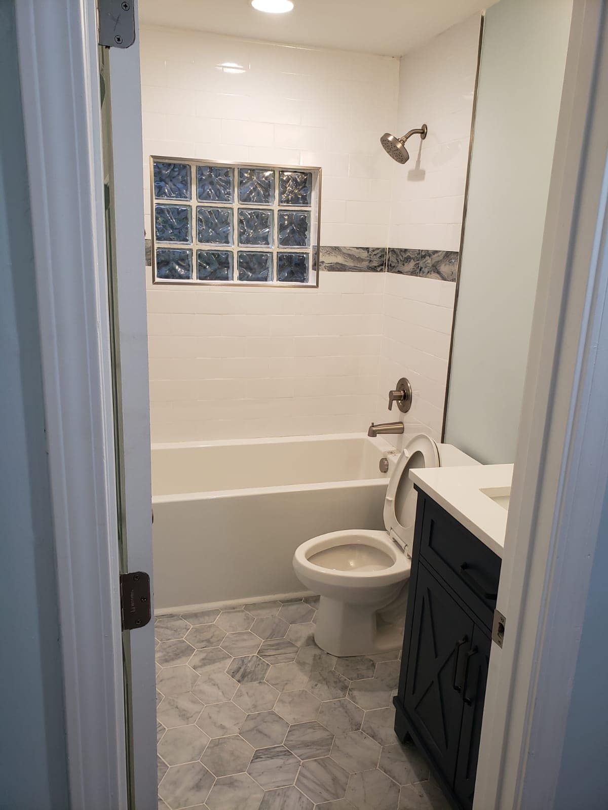Bathroom with white tile, a bathtub, and a toilet. Dark blue vanity on the right, glass block window.