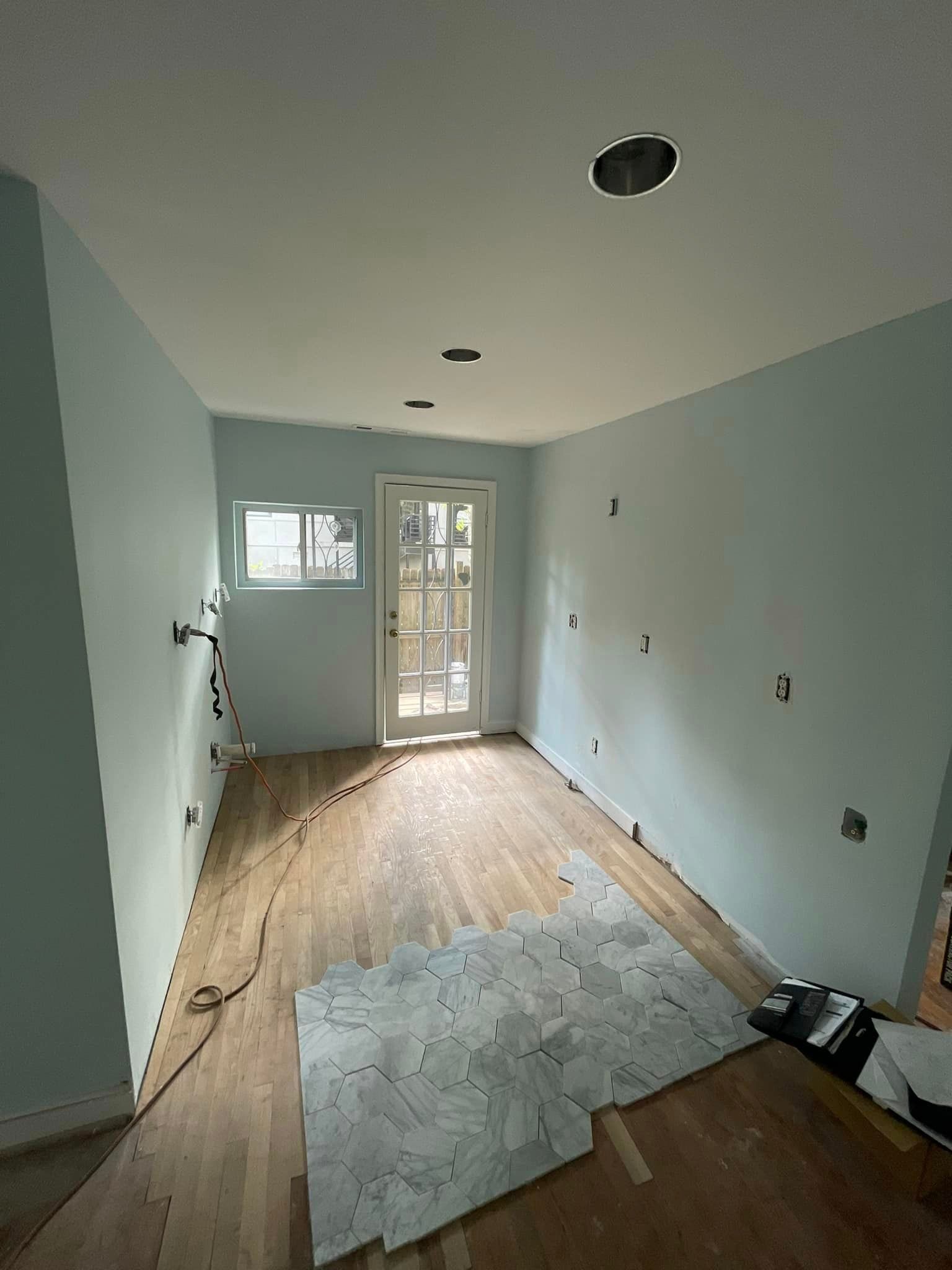 Room with light blue walls, wood floor, door, window, and ceiling lights, under renovation.