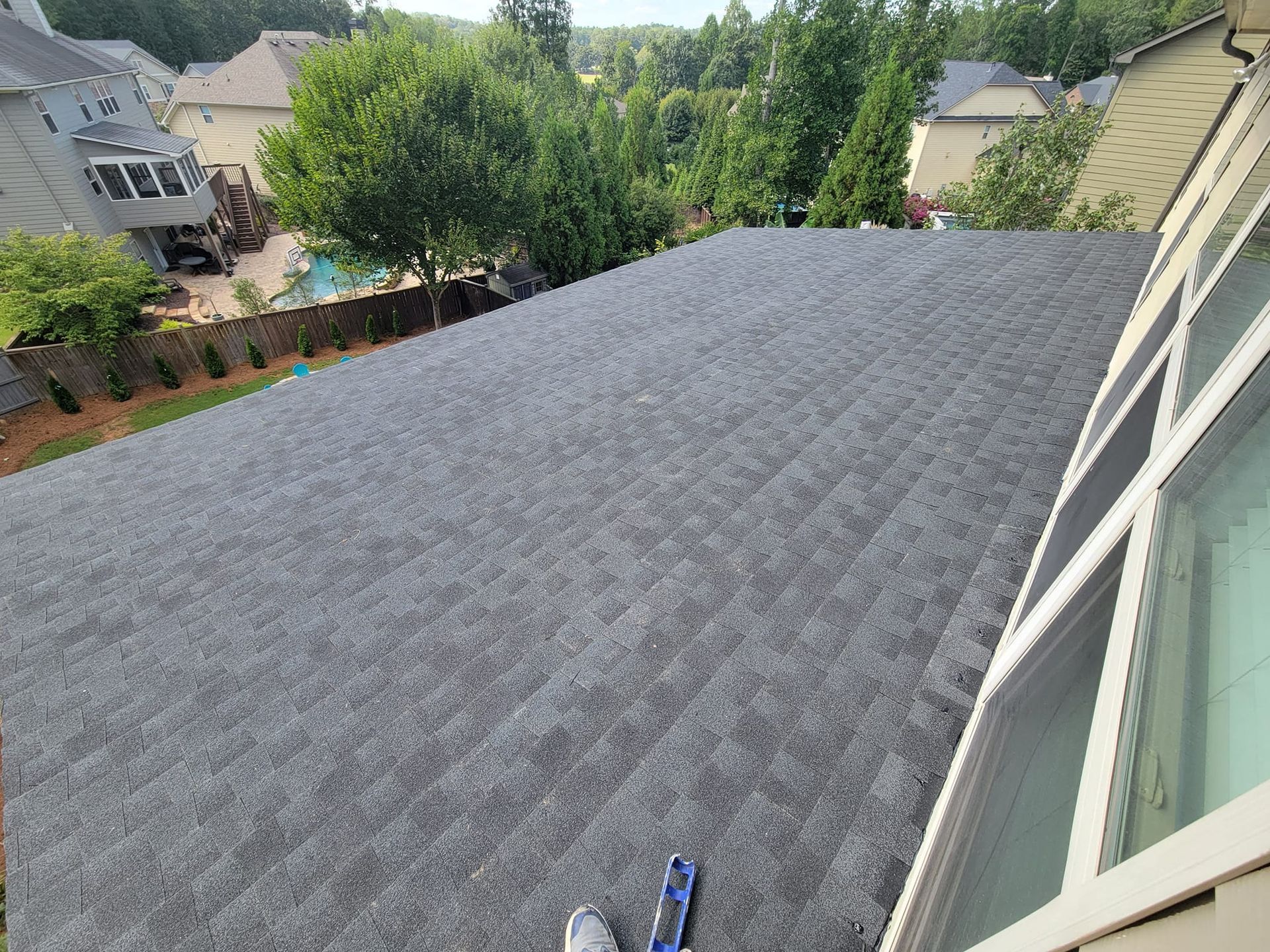 A gray asphalt shingle roof, viewed from above, with a window and trees in the background.