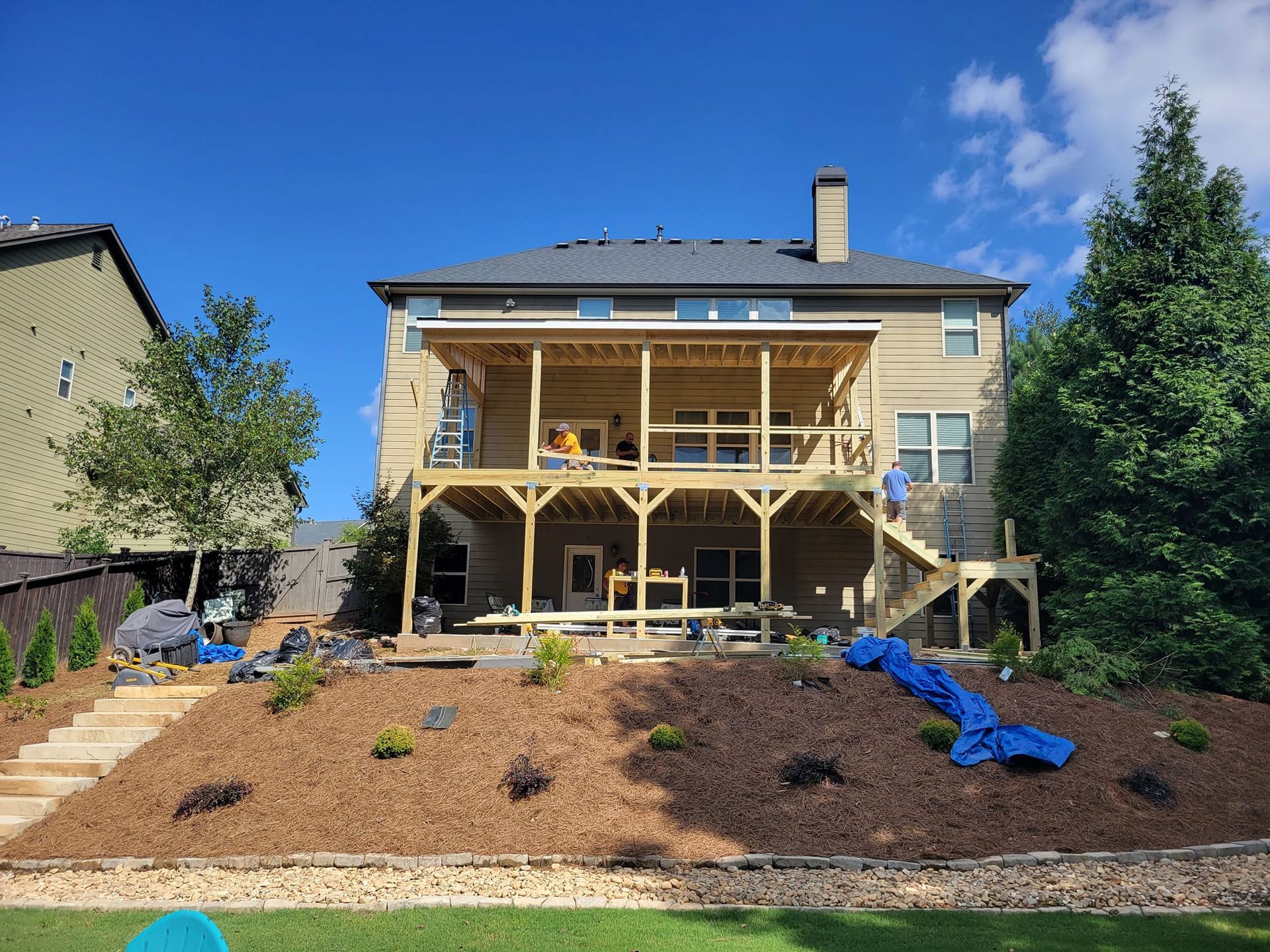 Two-story house with newly built wooden deck; workers on deck, landscaping in front, blue sky.
