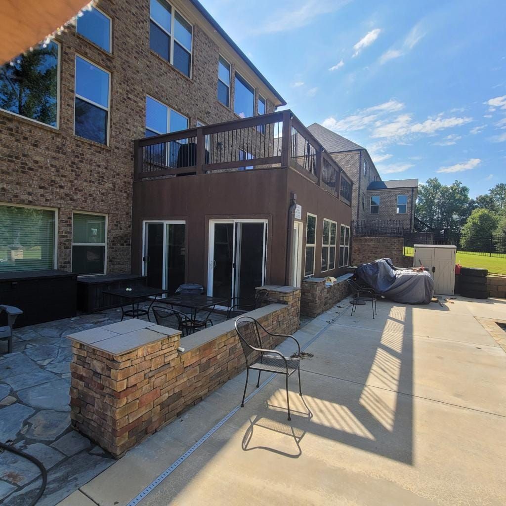 Back of a two-story brick house with a brown deck and patio. Stone wall and sunny blue sky.