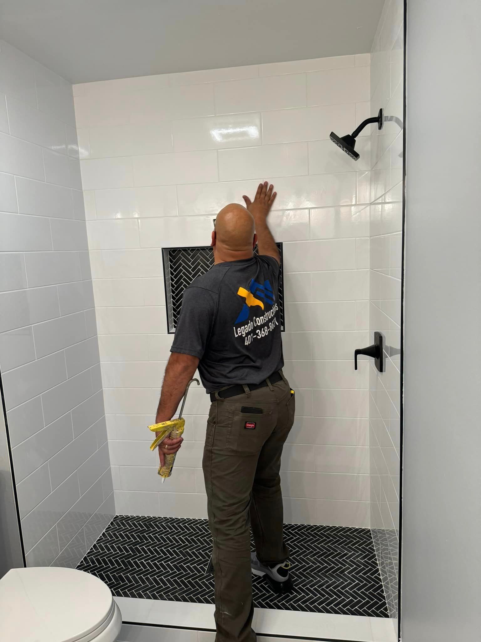 Man installing tile in a shower, reaching for the wall. White tiled walls with black fixtures.