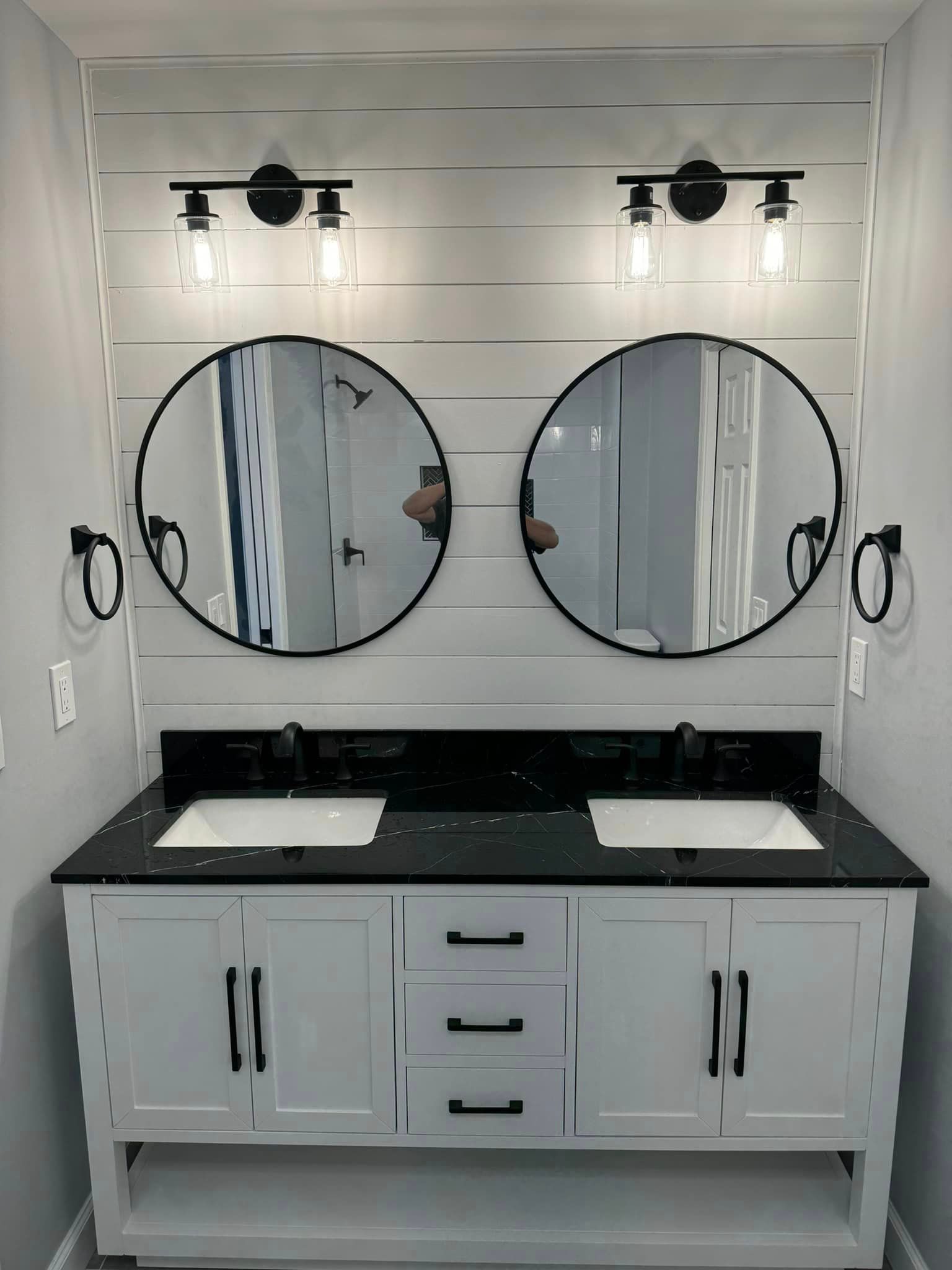 Bathroom vanity with two round mirrors, black countertops, and white cabinets.