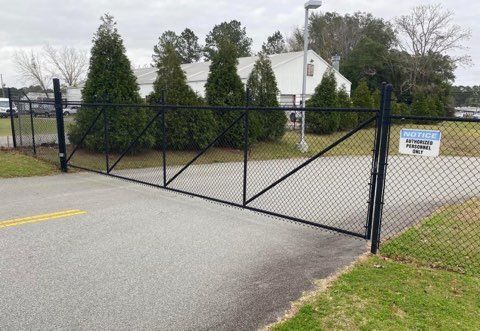 Black chain-link fence gate on paved surface; building and trees in background. 