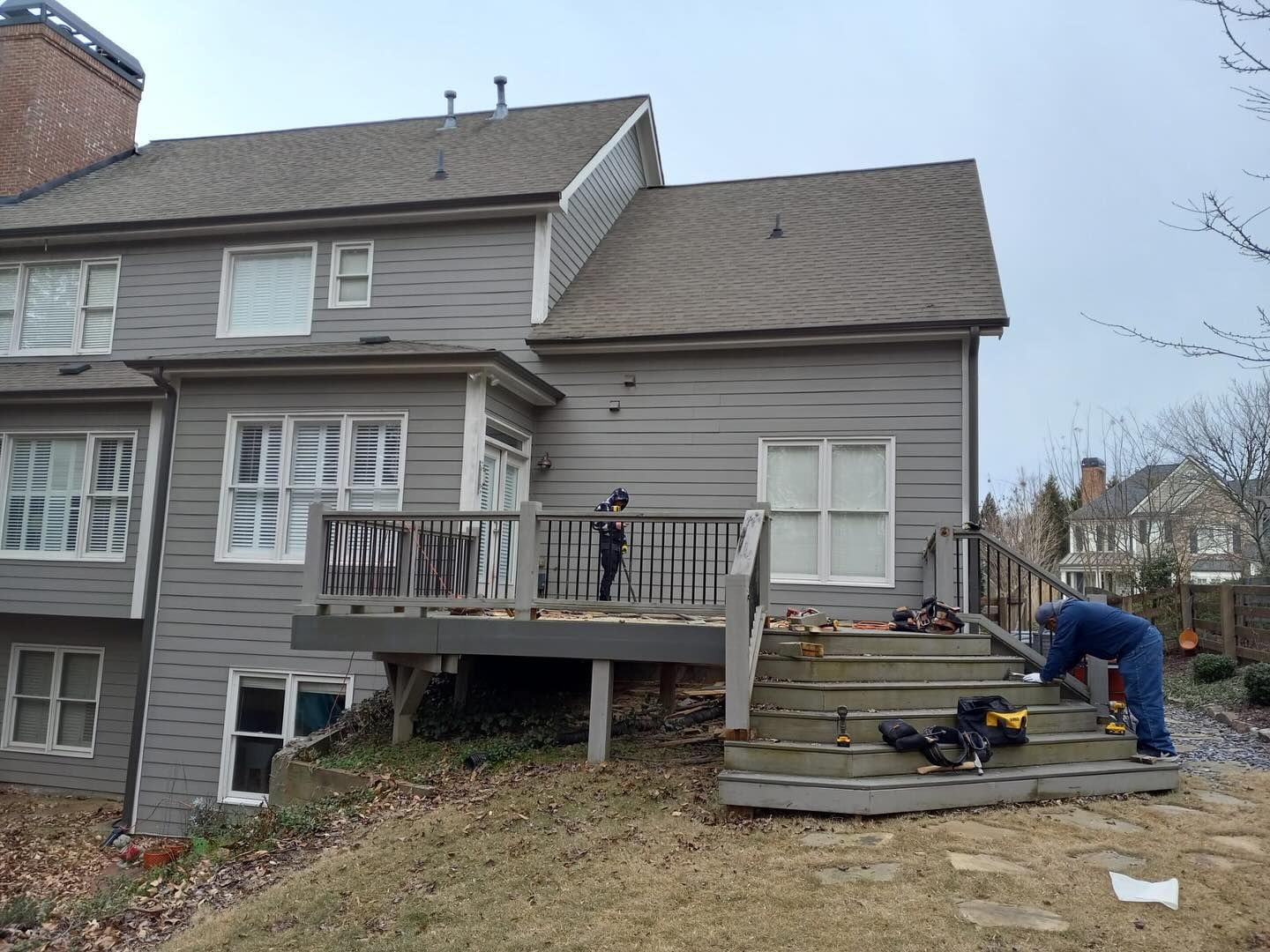 Workers on a gray house deck during construction. One works on steps, another on the deck. Overcast day.
