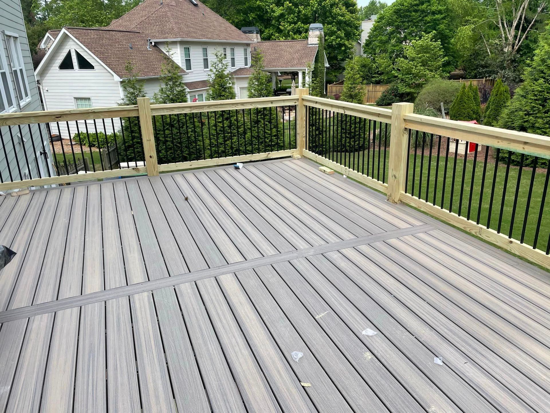 Wooden deck with gray planks and black railing, overlooking a yard with trees and a house.