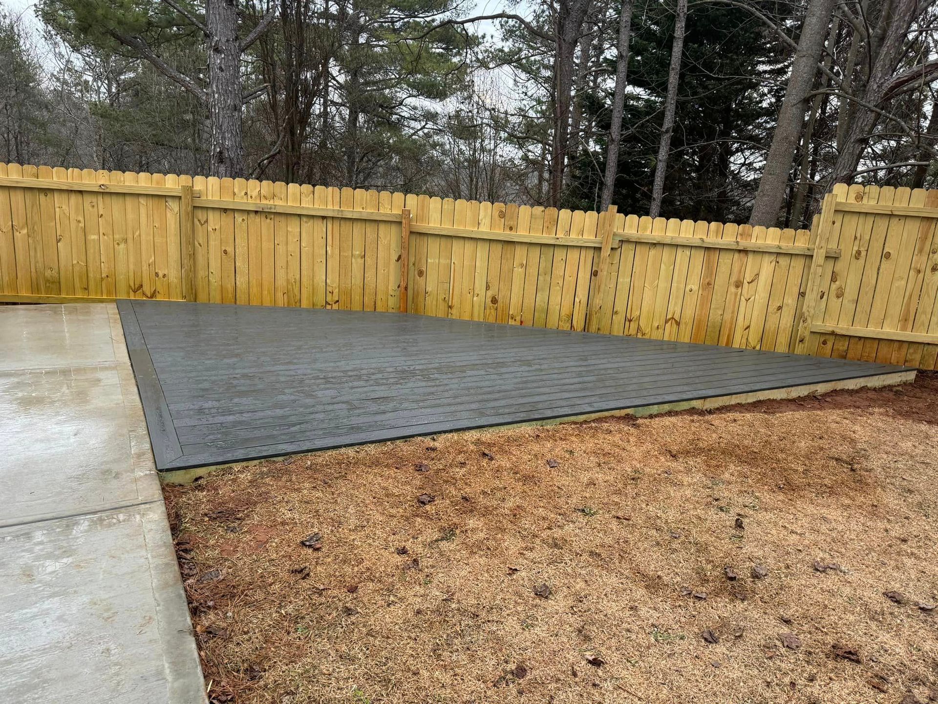 A rectangular, dark-colored patio platform in a backyard. A wooden fence surrounds it.