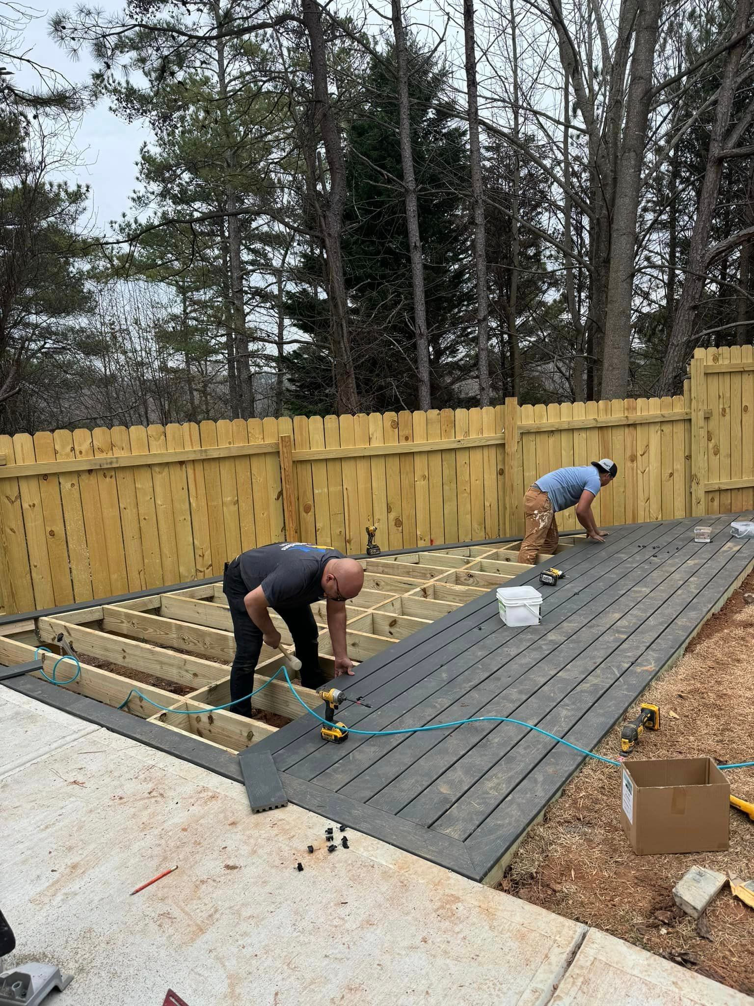 Two people constructing a wooden deck with dark gray boards. Construction site outdoors with a wooden fence.