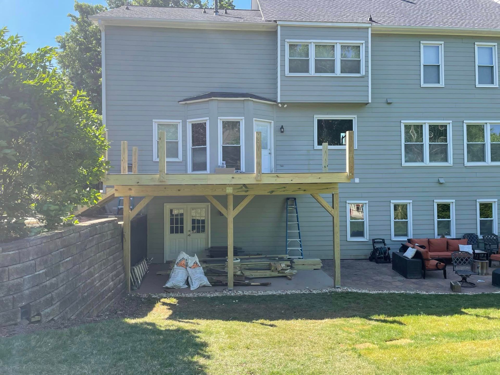 A two-story house with a wooden deck under construction; a patio, retaining wall, and green lawn.