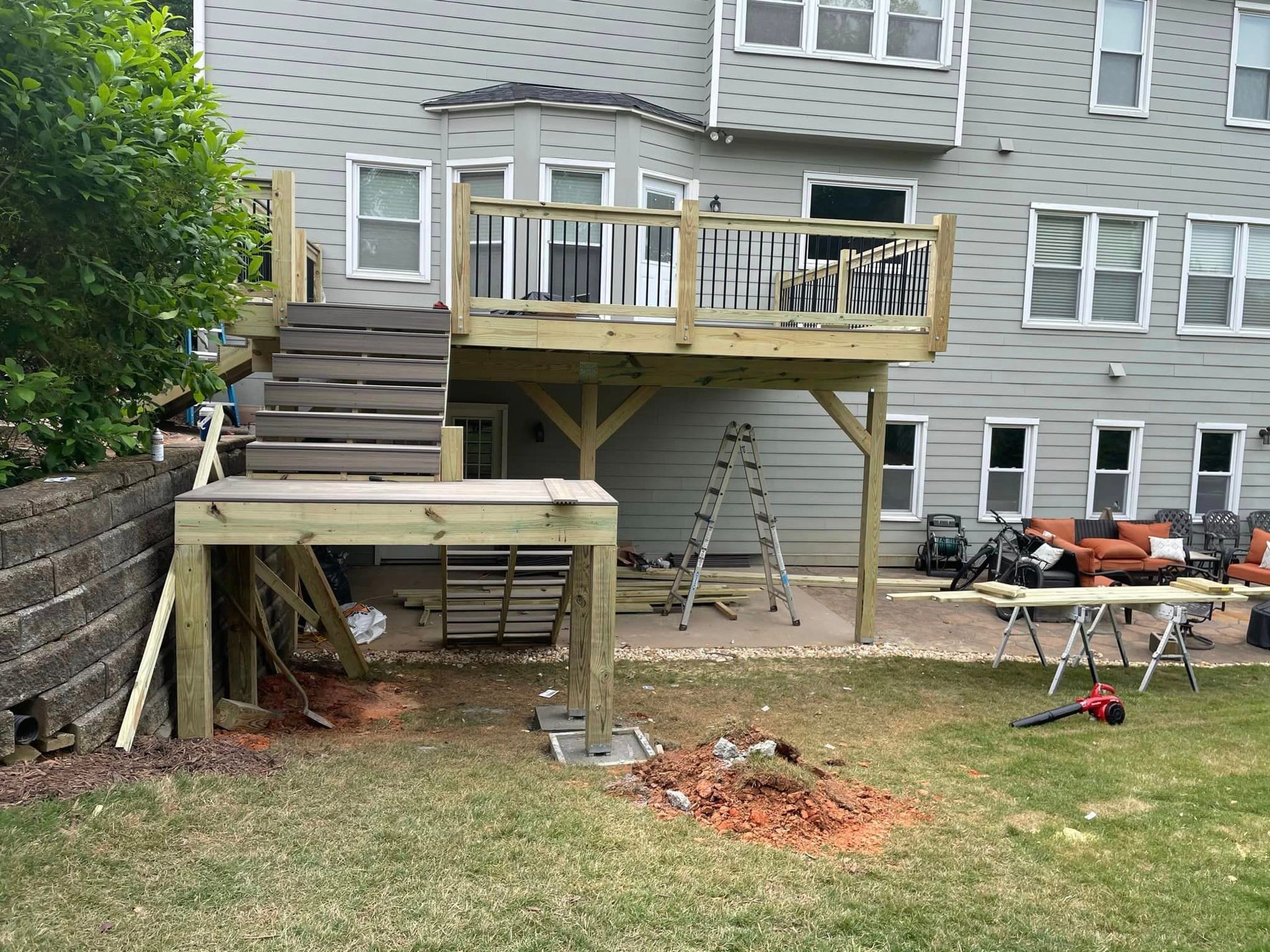 Wooden deck under construction attached to a two-story house. Lawn in foreground with landscaping and tools.