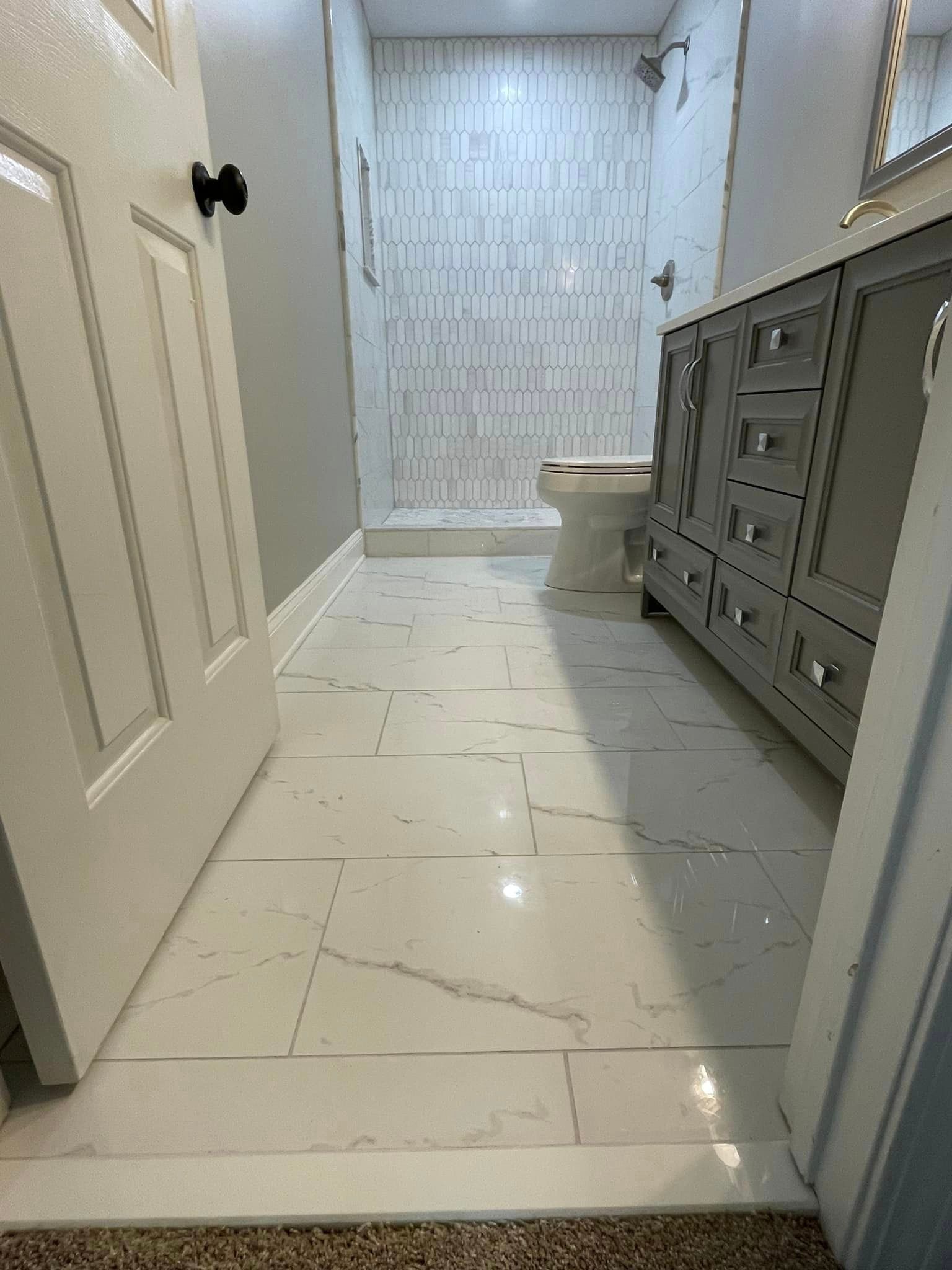 Bathroom with white tile floor, grey vanity, shower and walls. Door on the left.