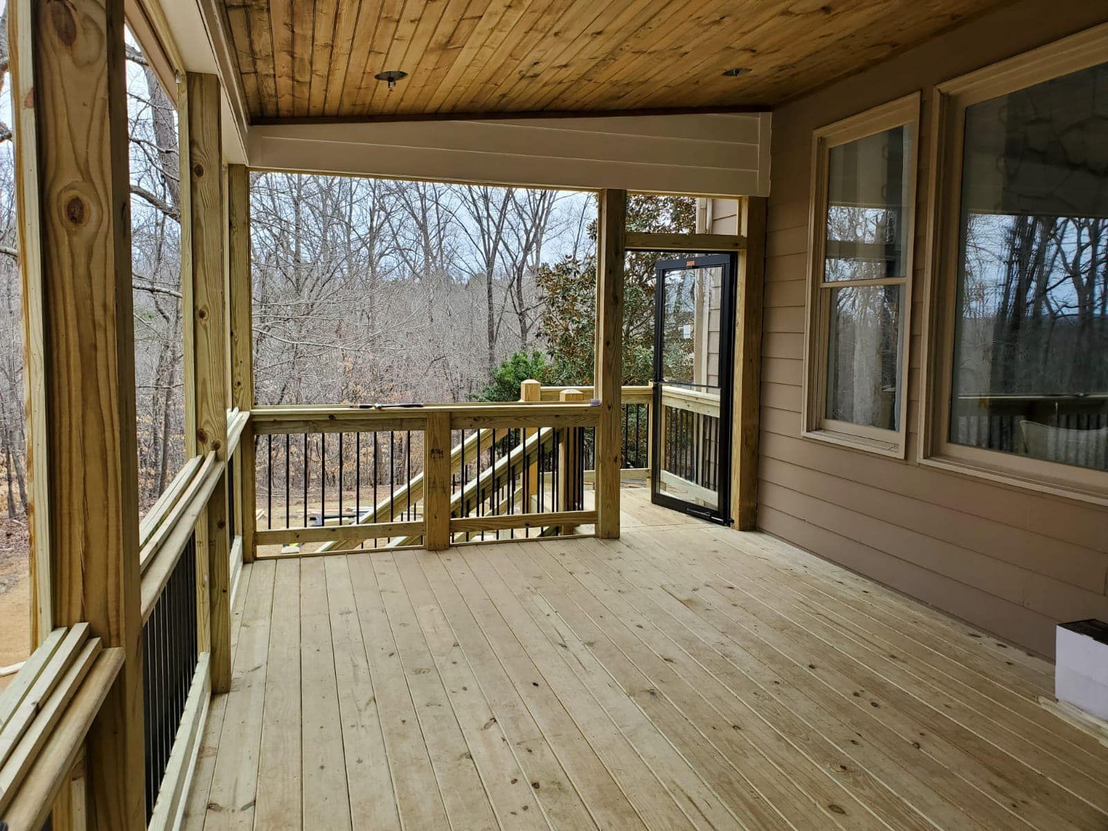 Wooden deck with railing, open doorway, and windows overlooking trees.