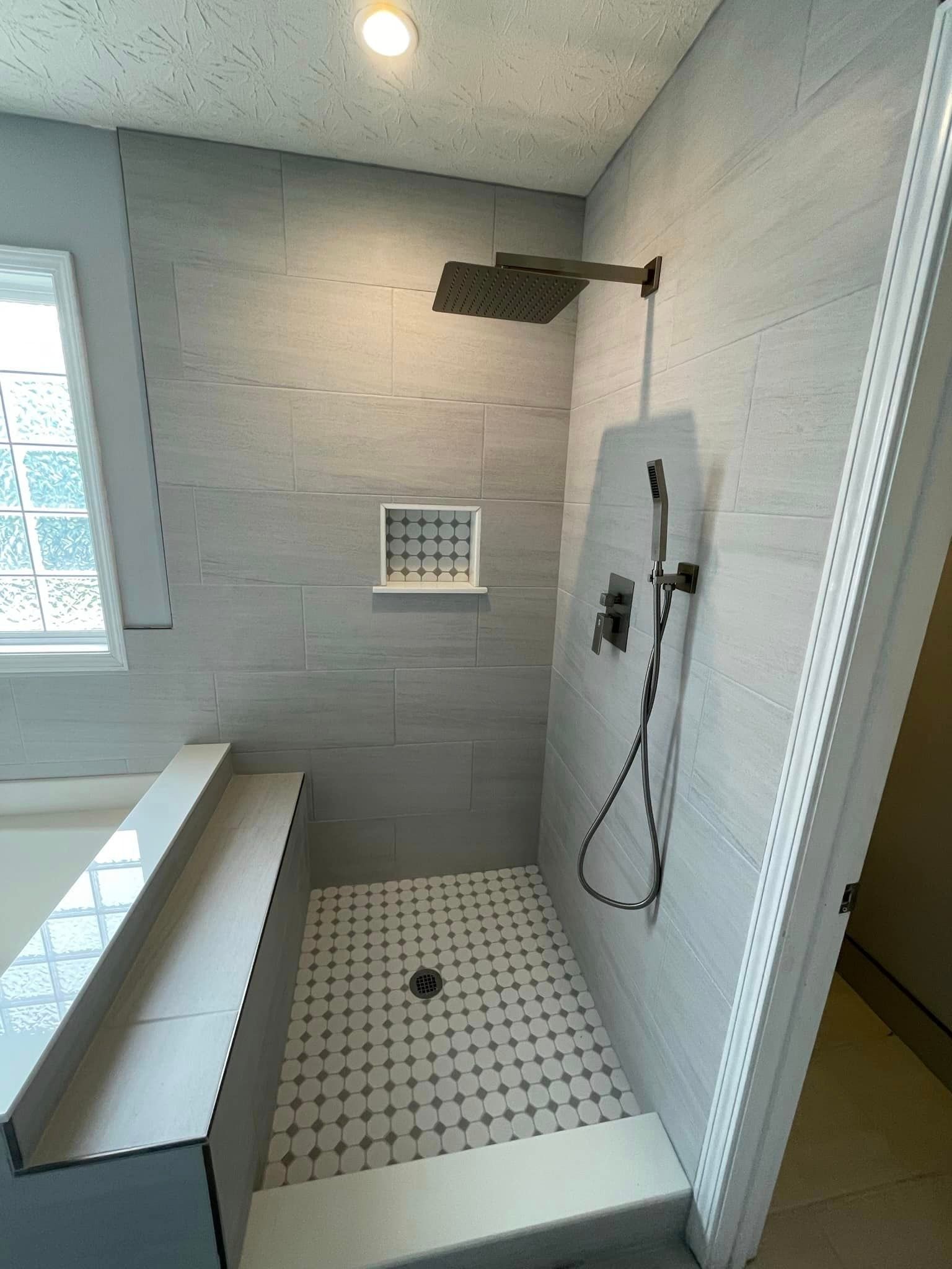 Bathroom with gray tiled shower, overhead showerhead, handheld shower, built-in bench, and patterned floor.