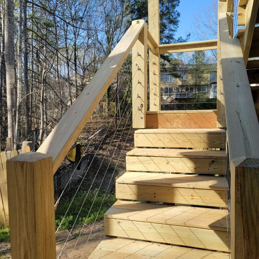 Wooden outdoor staircase with cable railings, leading to a house in a wooded area.