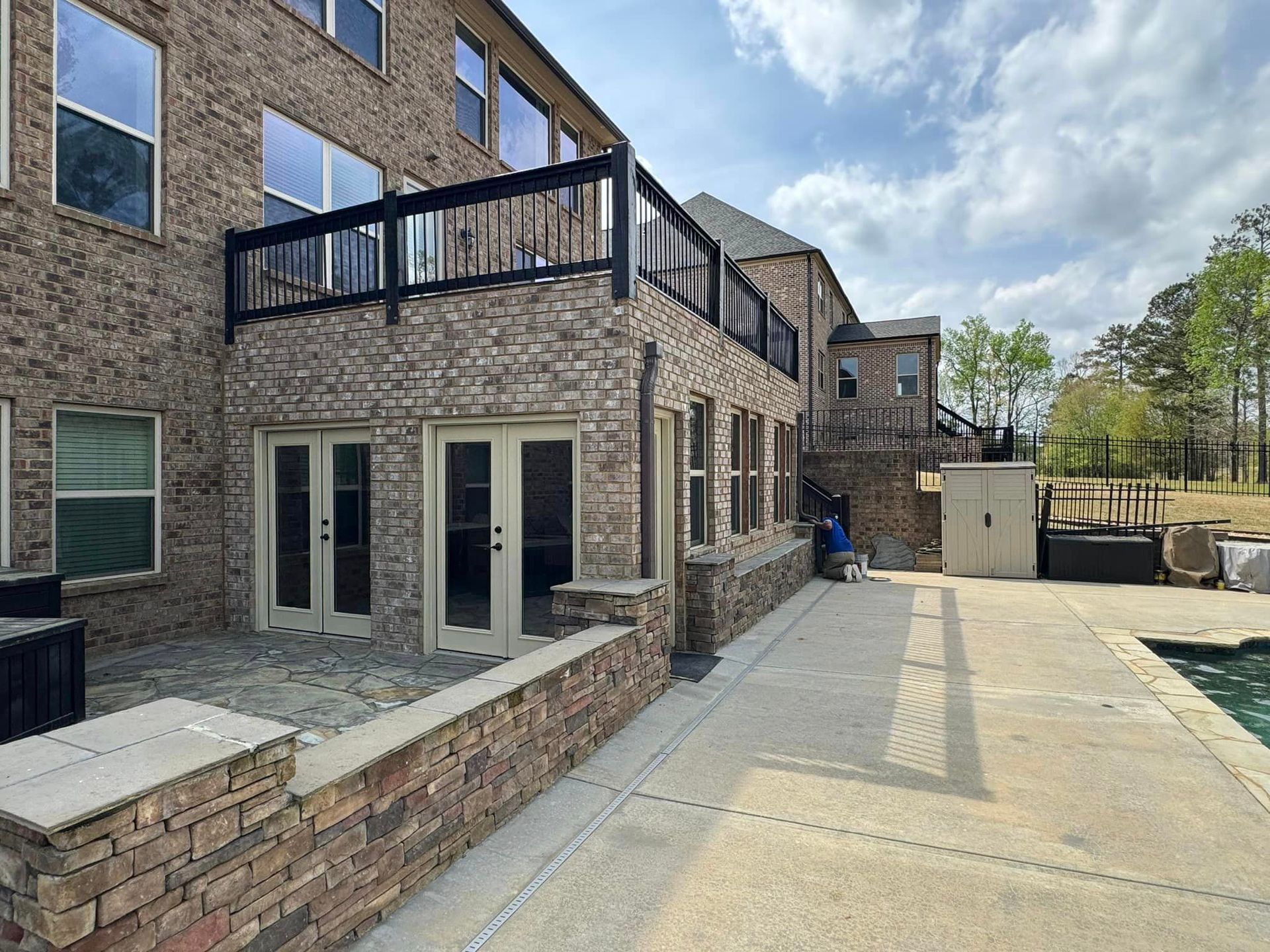 Brick house with deck, patio, and pool. Brown brick, black railing, blue sky.