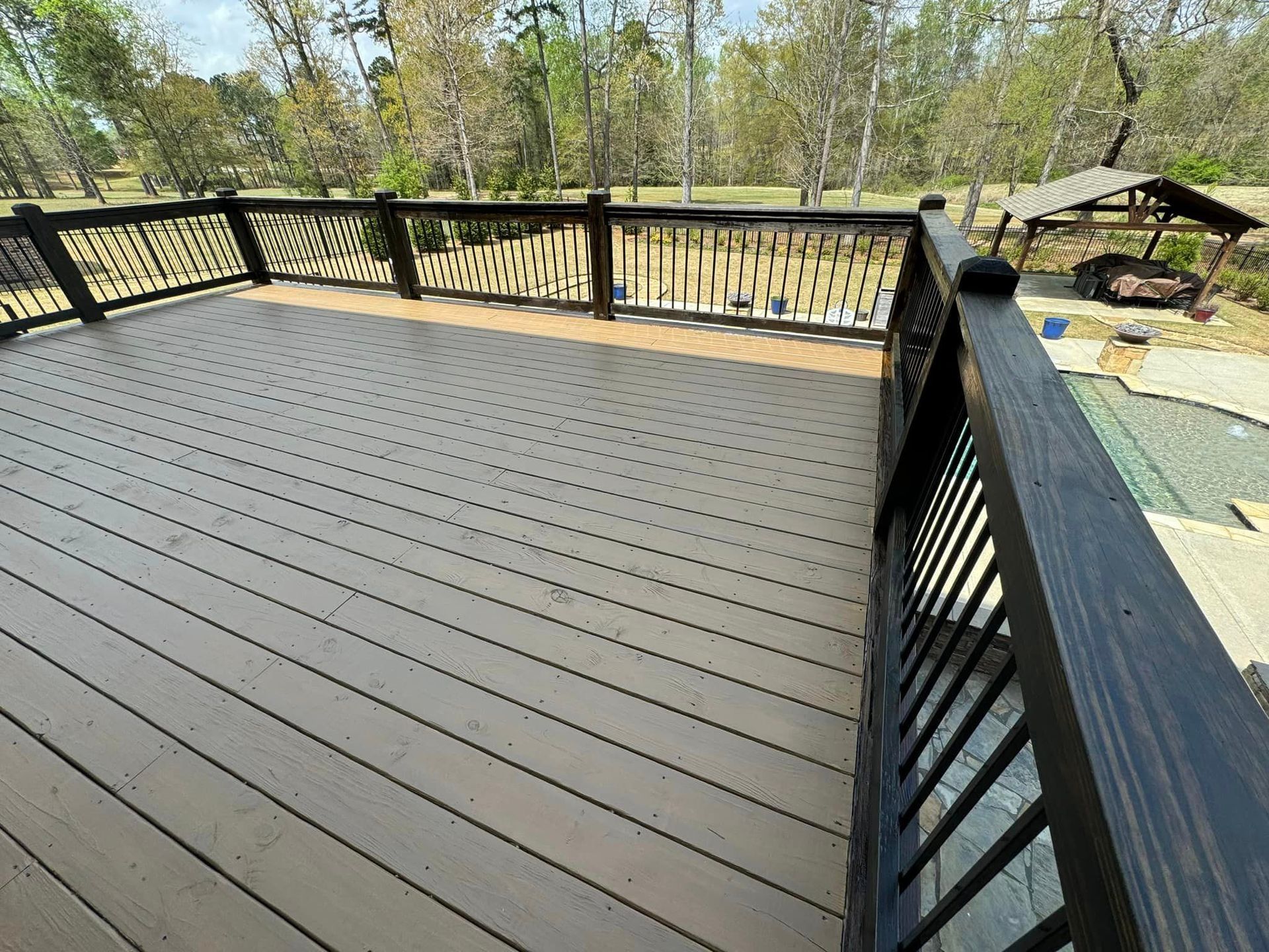 Wooden deck with black railings overlooking a backyard with trees and a small shelter.