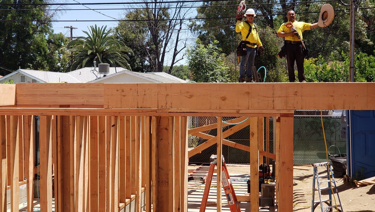 Construction workers atop a wooden structure, waving arms. Sunny outdoor setting with trees and houses.