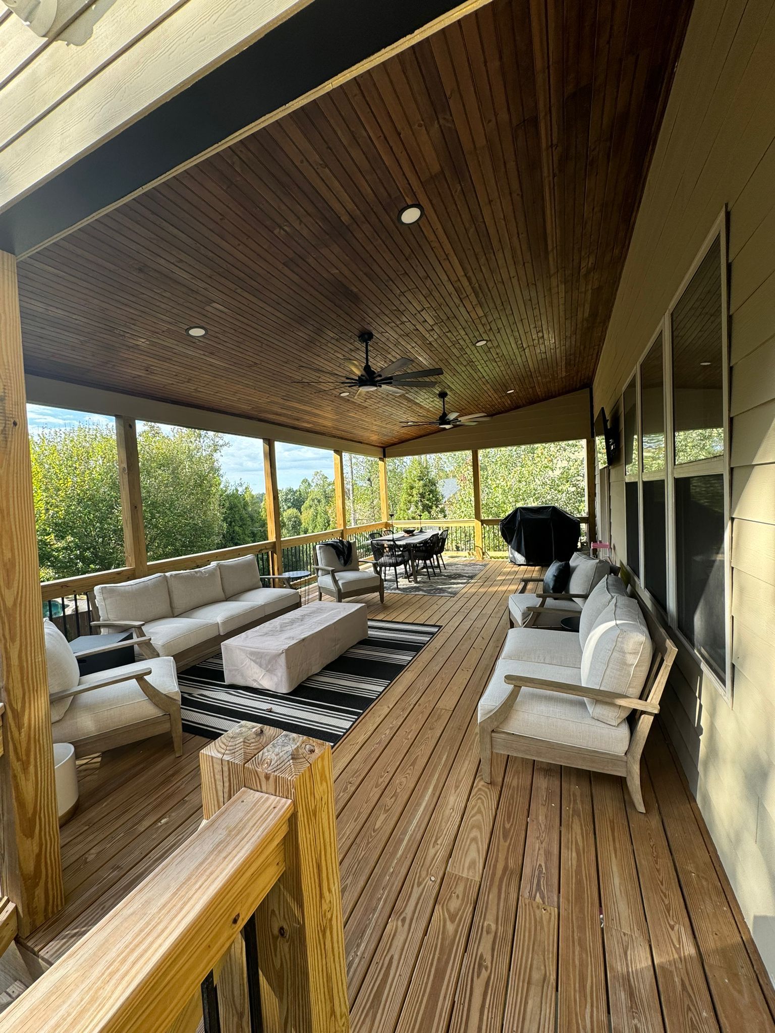 Covered wooden porch with seating area; beige sofas, black and white rug, grill, and ceiling fans.