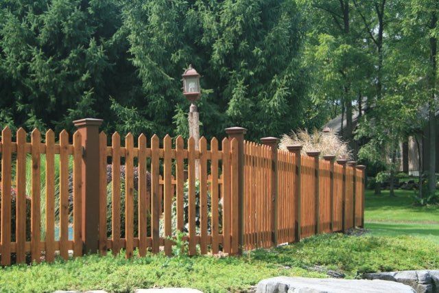 Brown wooden picket fence with trees and grass in a sunny outdoor setting.