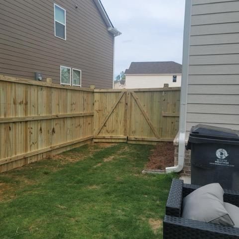 Wooden fence encloses a backyard with green grass, a gate, and a black trash can.