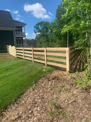 Wooden fence bordering a grassy yard and wooded area under a blue sky.