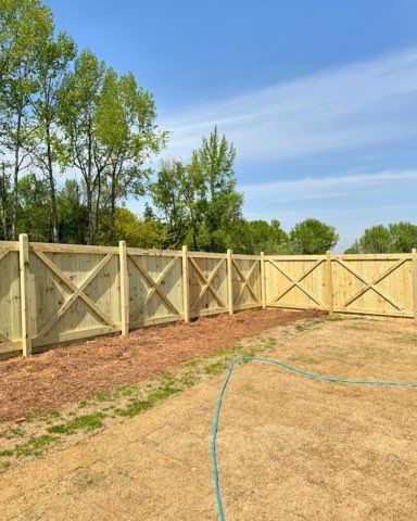 Wooden fence with X-shaped crossbeams encloses a yard, with a clear blue sky and trees in the background.