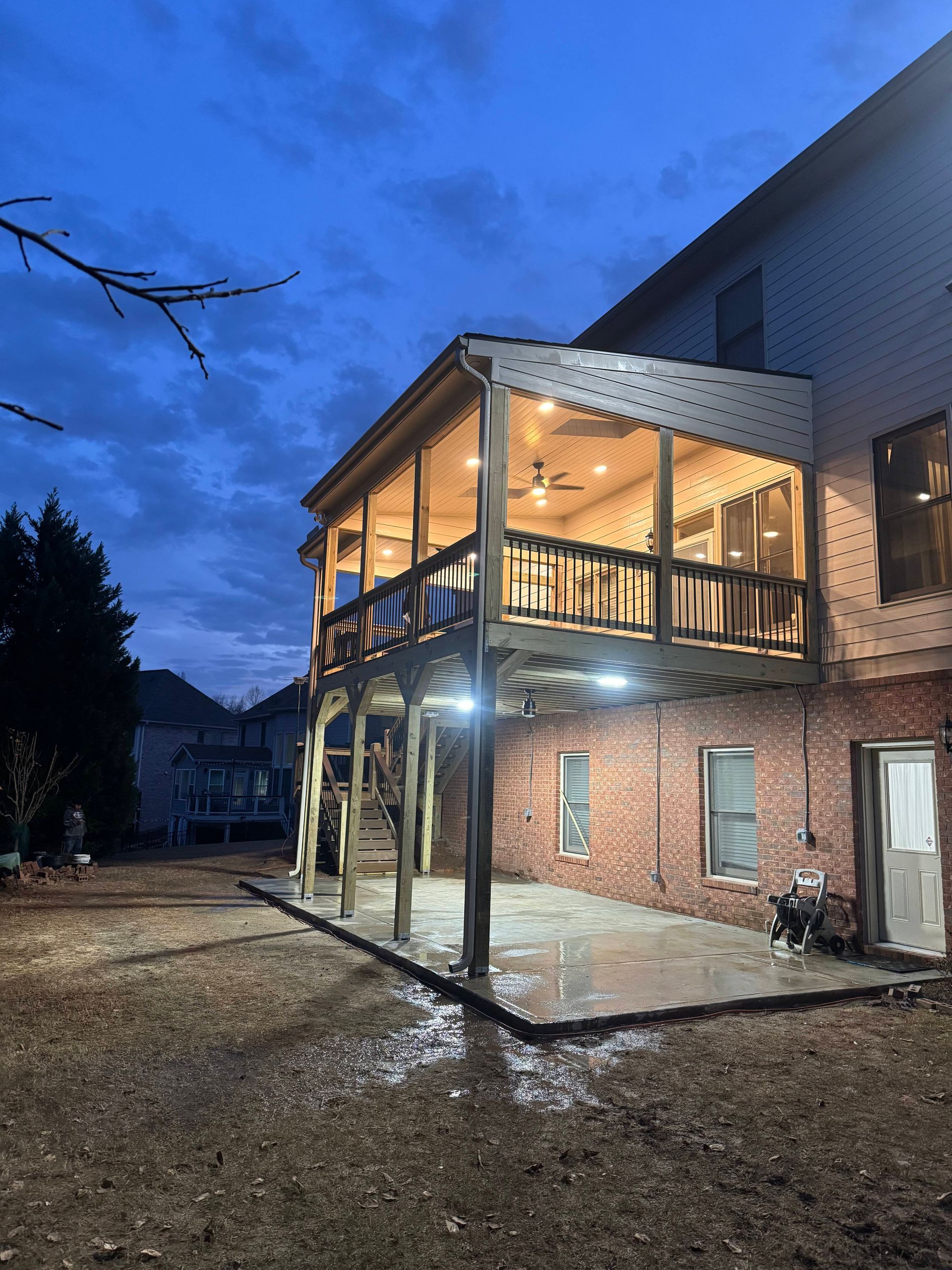 Two-story deck with lights on, attached to a brick home. Evening, wet ground.