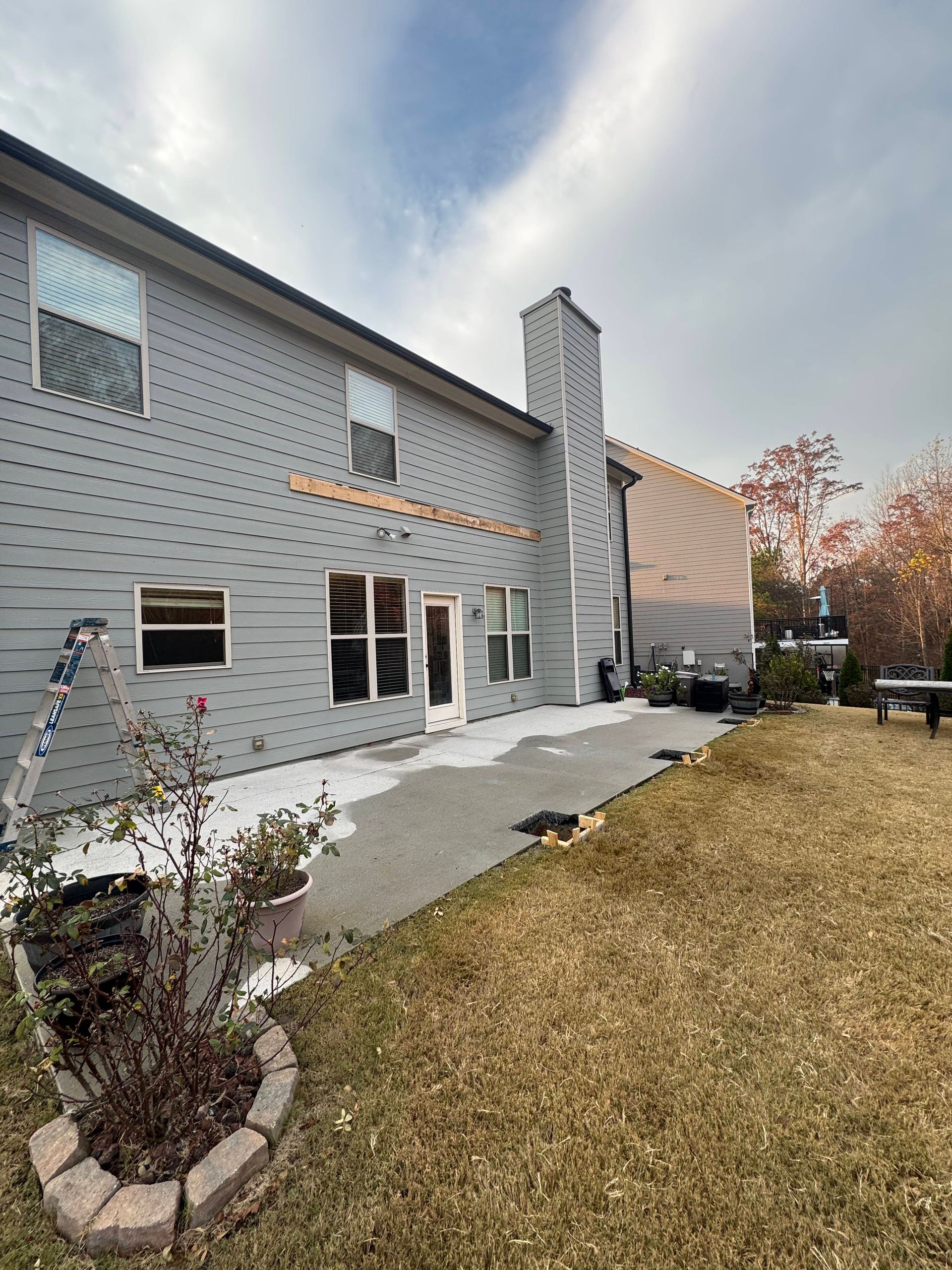 Backyard view of a gray house, chimney, concrete patio, and brown grass under a cloudy sky.