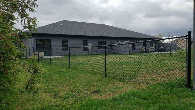 A gray house with a black chain-link fence in front, overcast sky, green lawn.
