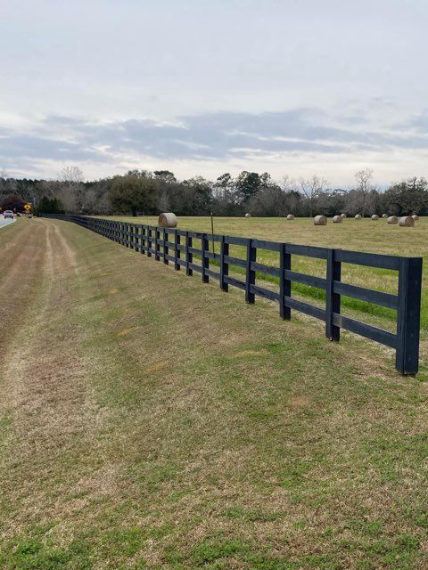 Black wooden fence in a grassy field, hay bales in the distance, overcast sky.