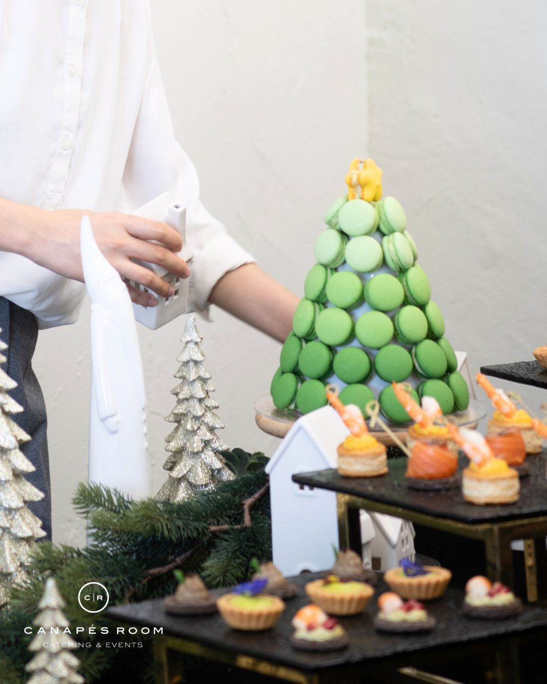 Person arranging macarons in the shape of a Christmas tree next to appetizers.
