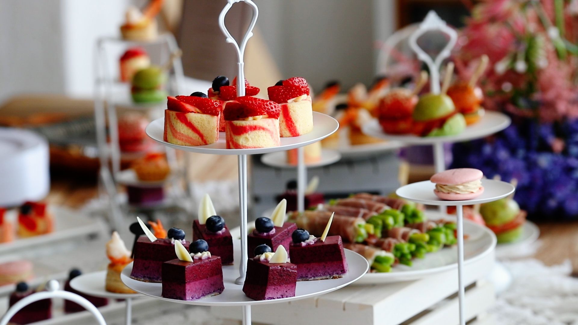 Tiered dessert tray with mini cakes, fruit tarts, and macarons in a bright setting.
