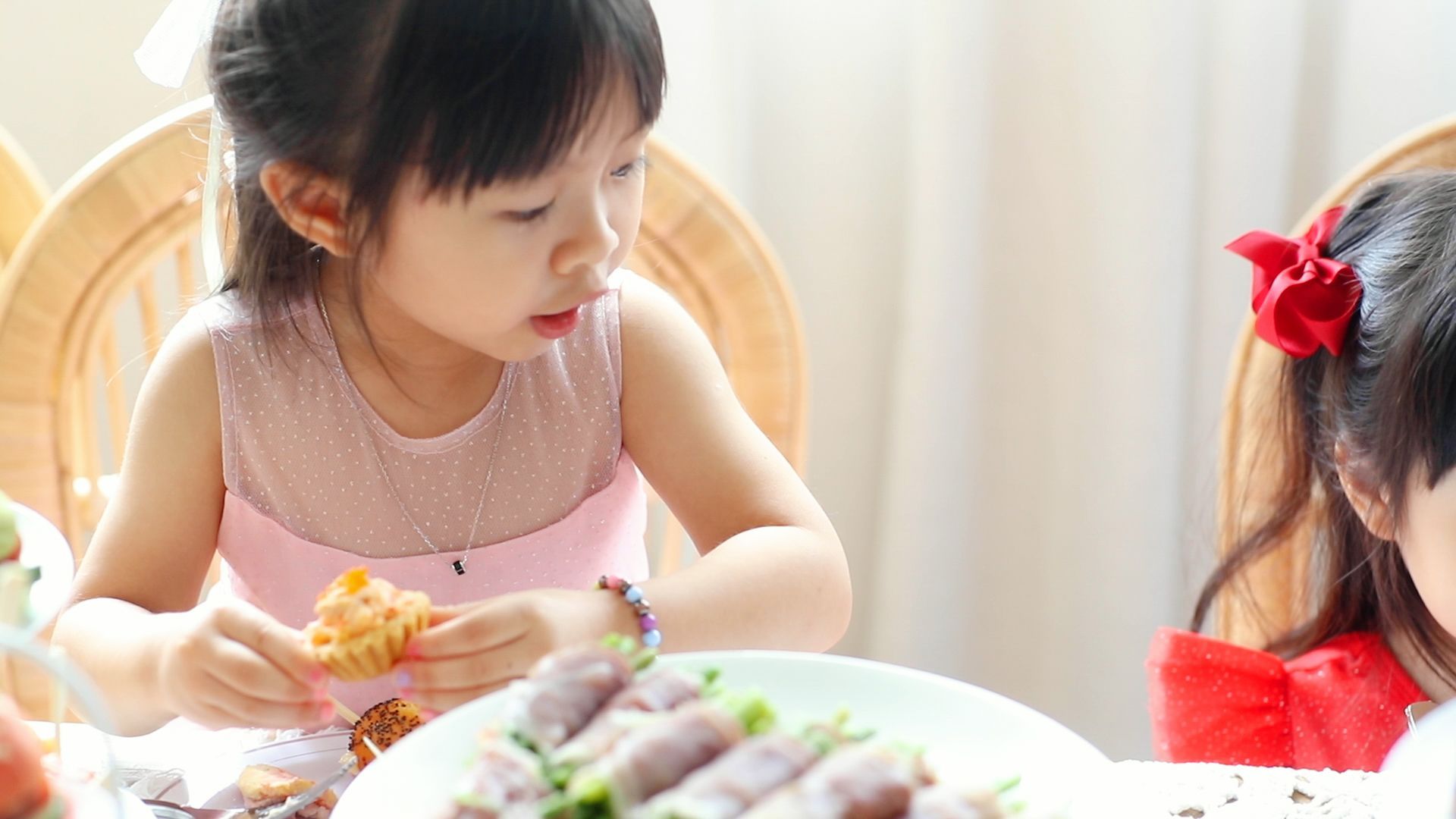 Two young children eating at a table; one in a pink dress, another with a red bow.