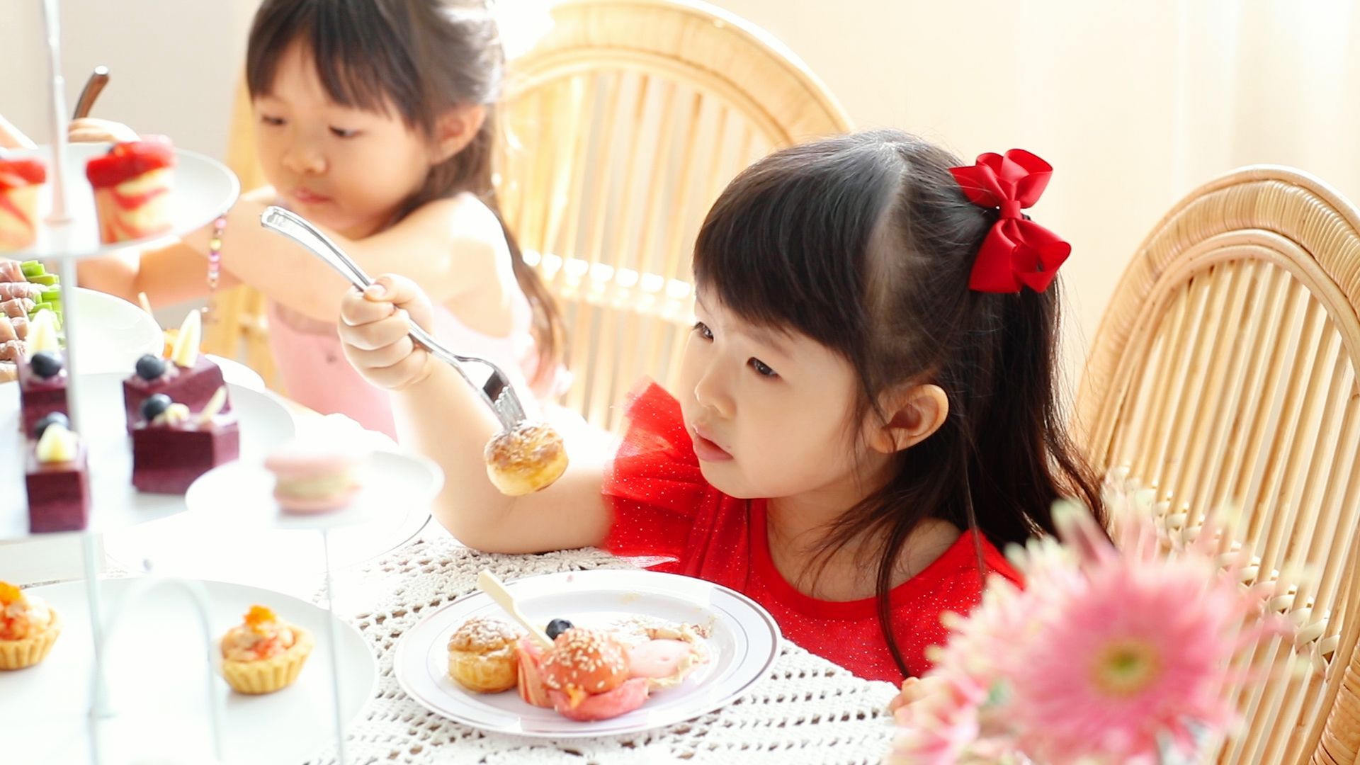 Two children at a table selecting treats from a dessert display. One child uses tongs; red accents.