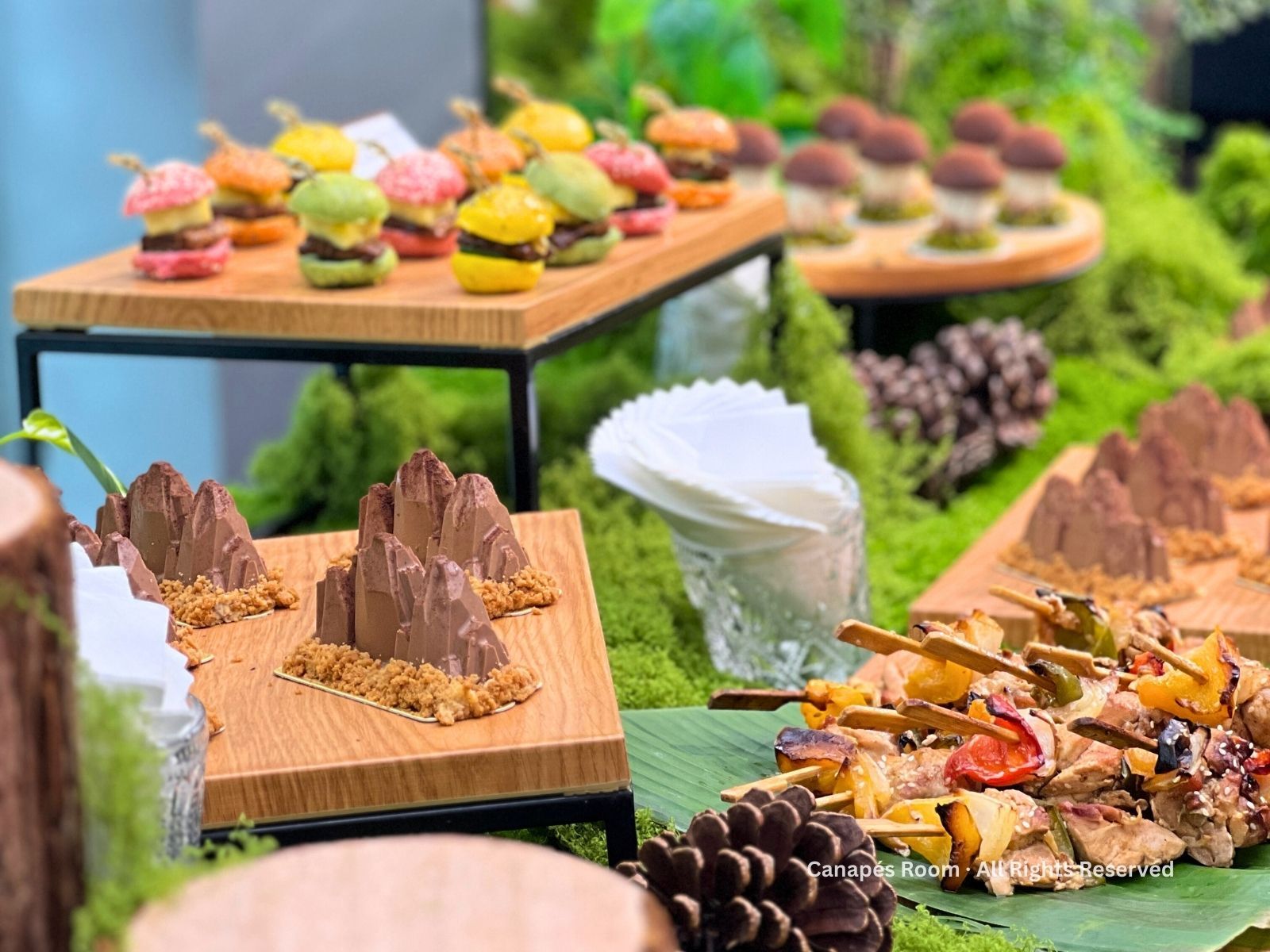 Appetizers on wooden platters: mini burgers, mountain-shaped chocolates, skewers, set on moss and greenery.