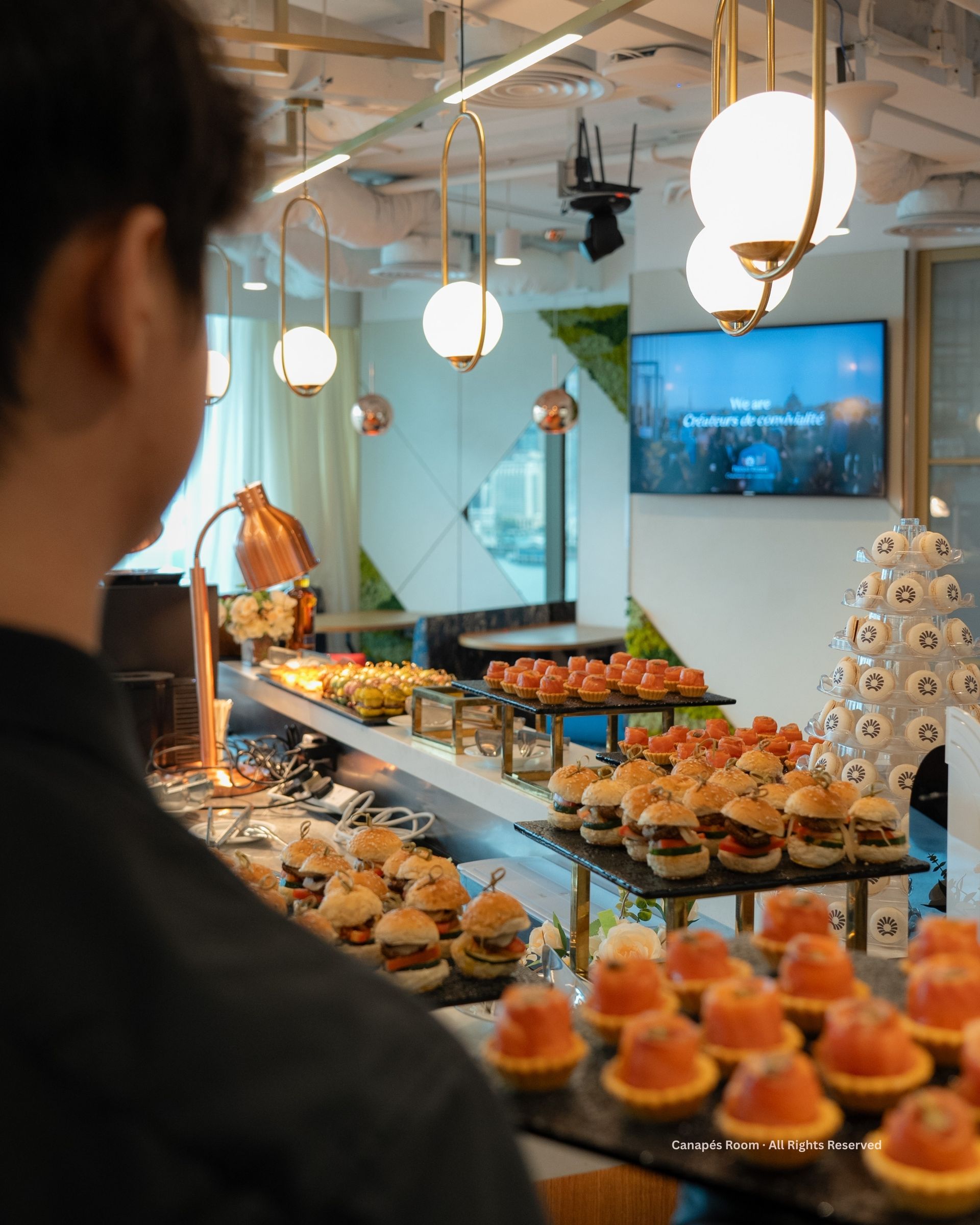 Person looking at a buffet of appetizers including sliders, and tarts under decorative lighting.