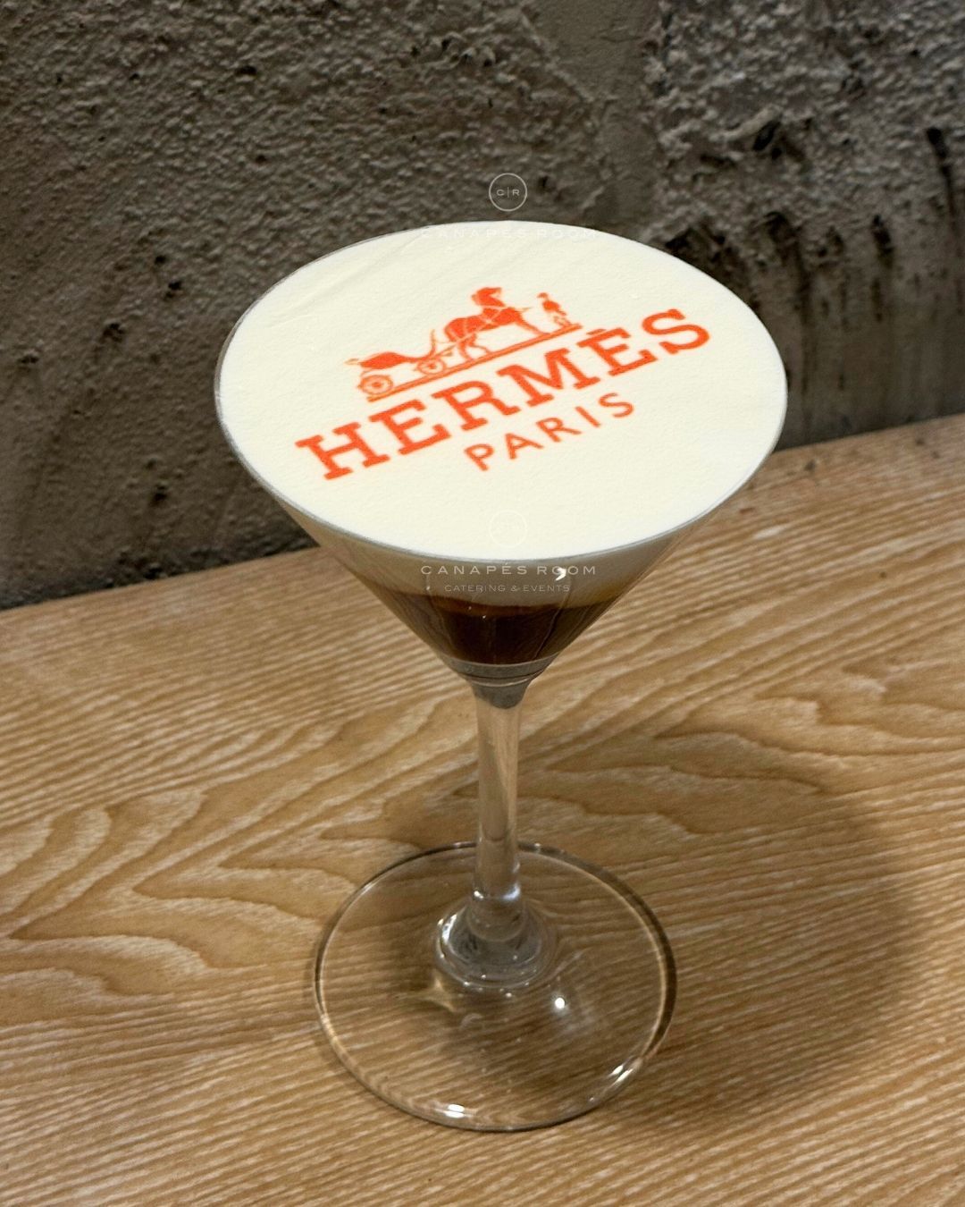 Cocktail in a glass with white foam top, the Hermes logo printed on it.