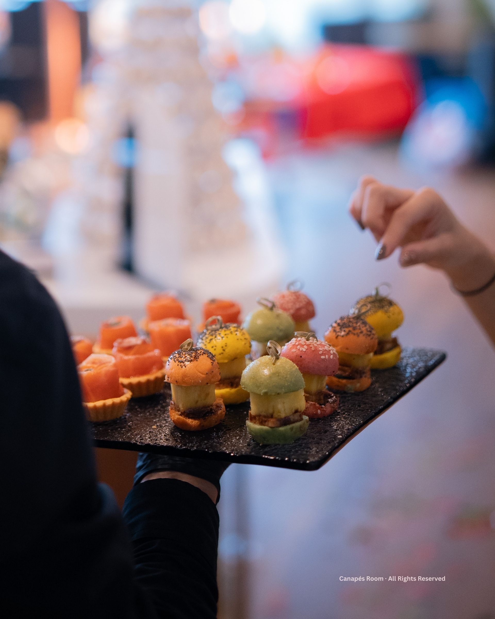 A person holds a tray of colorful appetizers, someone reaches to take one. Blurred background.
