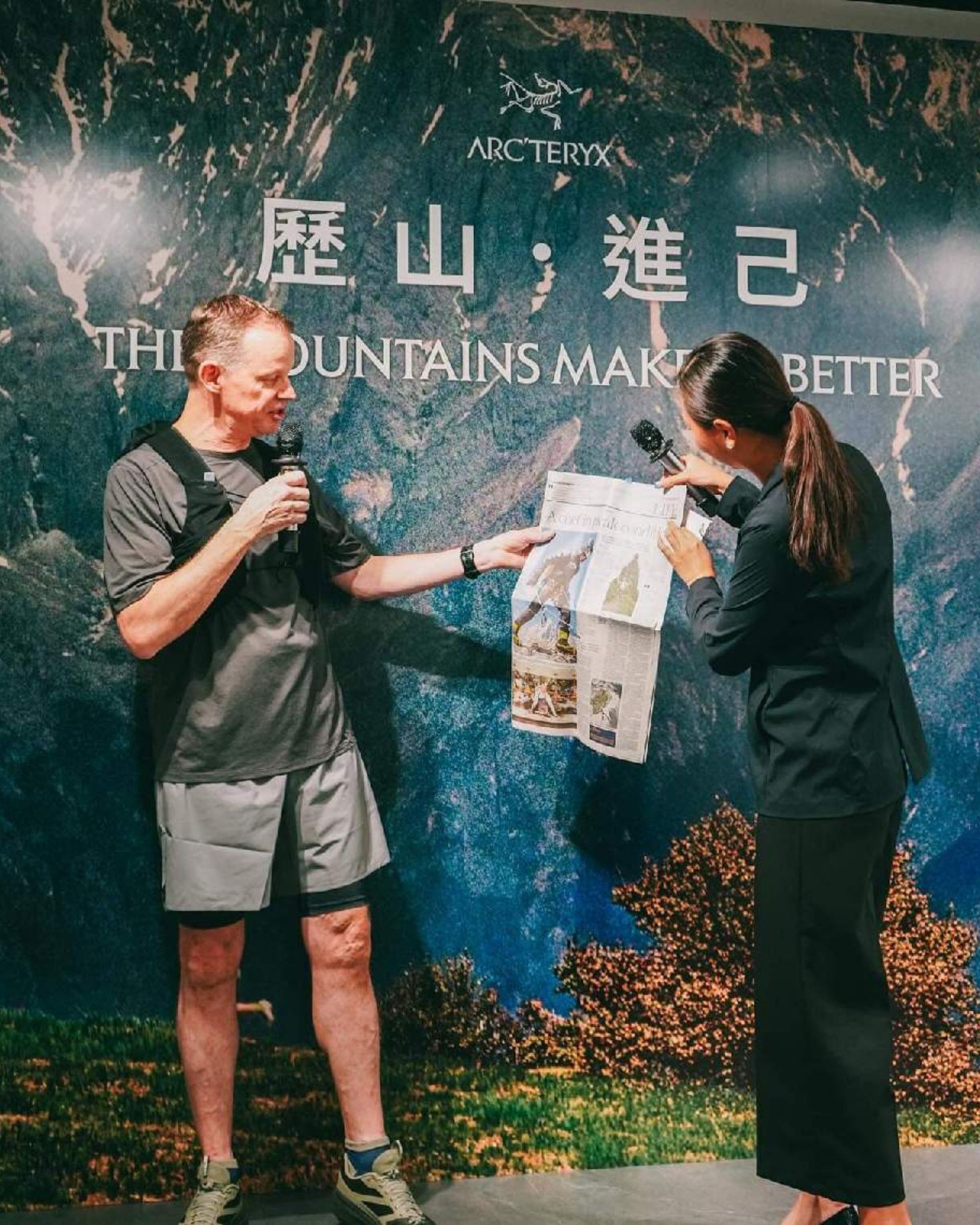 Man and woman at event, man holding a printed item, woman pointing to it. Blue backdrop with text.